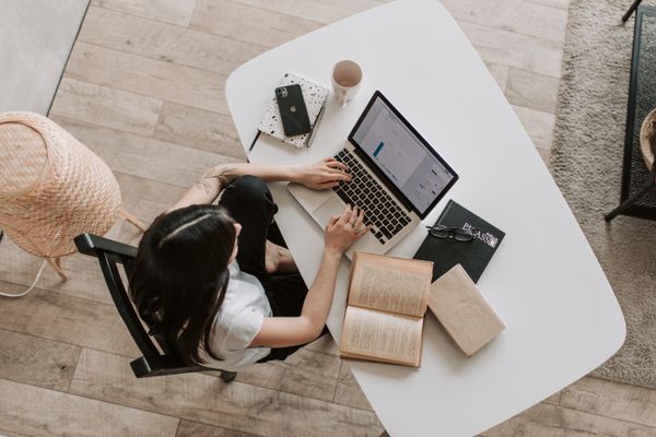 Overhead view of a woman working on a laptop at a bright home desk with books, phone and coffee